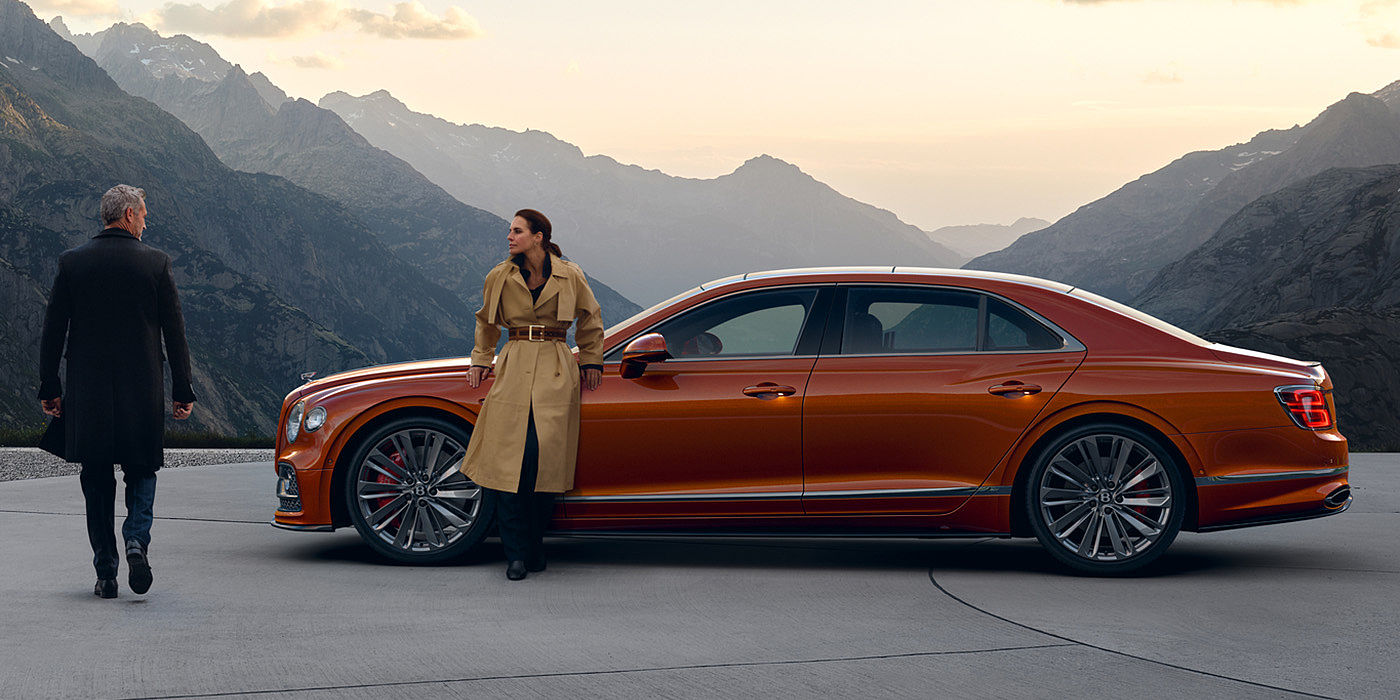 Bentley Brussels Bentley Flying Spur Speed parked in Orange Flame coloured exterior parked, with mountainous background and two people in view.
