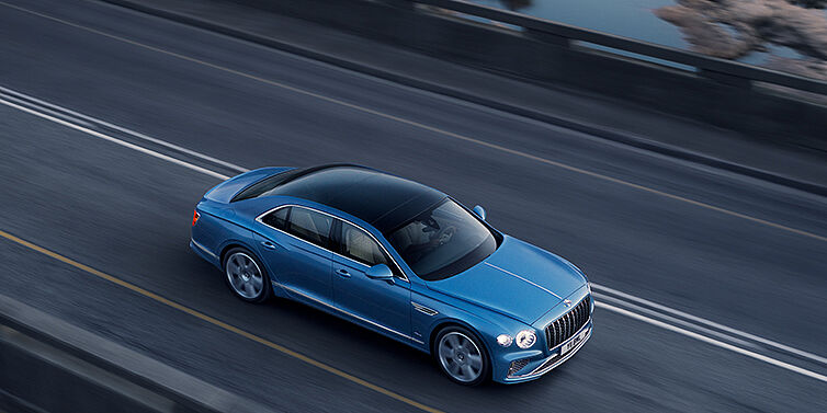Bentley Brussels New Bentley Flying Spur Azure sedan in Light windsor blue paint by Mulliner, with 22 inch Azure wheels seen from overhead driving dynamically on a country road