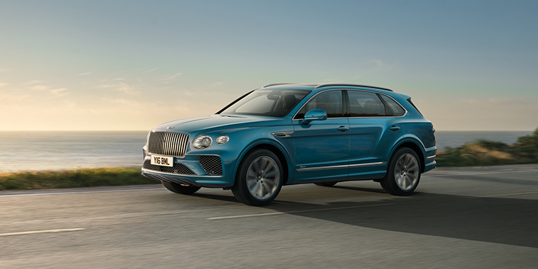 Bentley Brussels Bentley Bentayga Azure SUV in Topaz blue paint driving dynamically by the ocean with 22 inch 10 spoke directional wheels