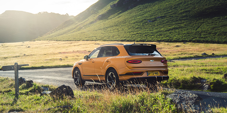 Bentley Brussels Bentley Bentayga S SUV rear three quarter in Sunburst Gold paint on a road surrounded by green hills