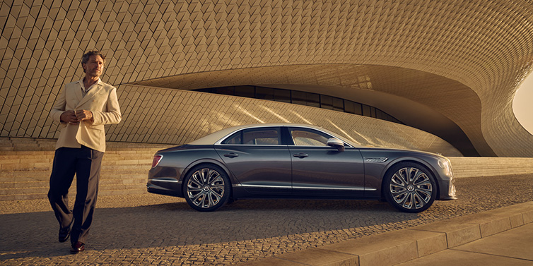 Bentley Brussels Bentley Flying Spur Mulliner sedan in Rose Gold over Magnetic duotone paint parked by a curved wall with person in suit jacket standing on the left